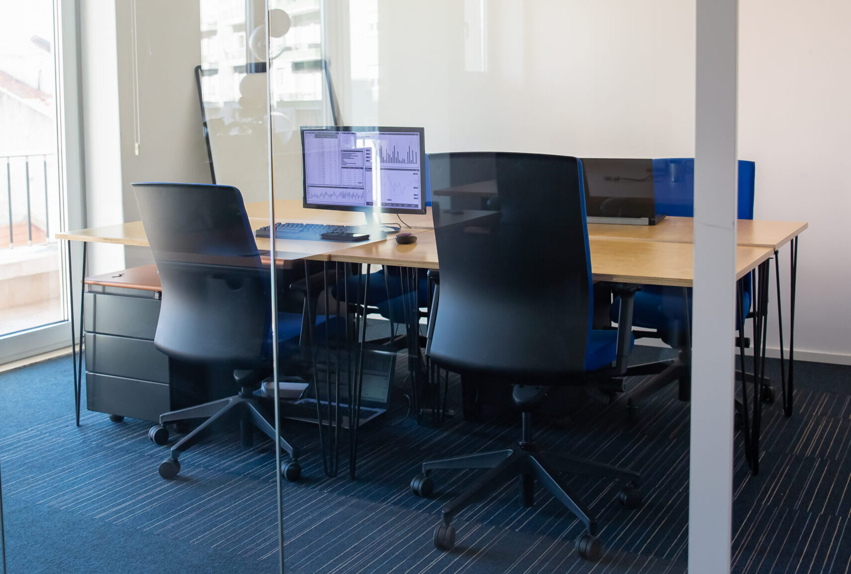 Empty boardroom behind glass wall. Meeting room with conference table, shared desk for team and workplaces. Trading graphs on monitor. Office interior or commercial real estate concept